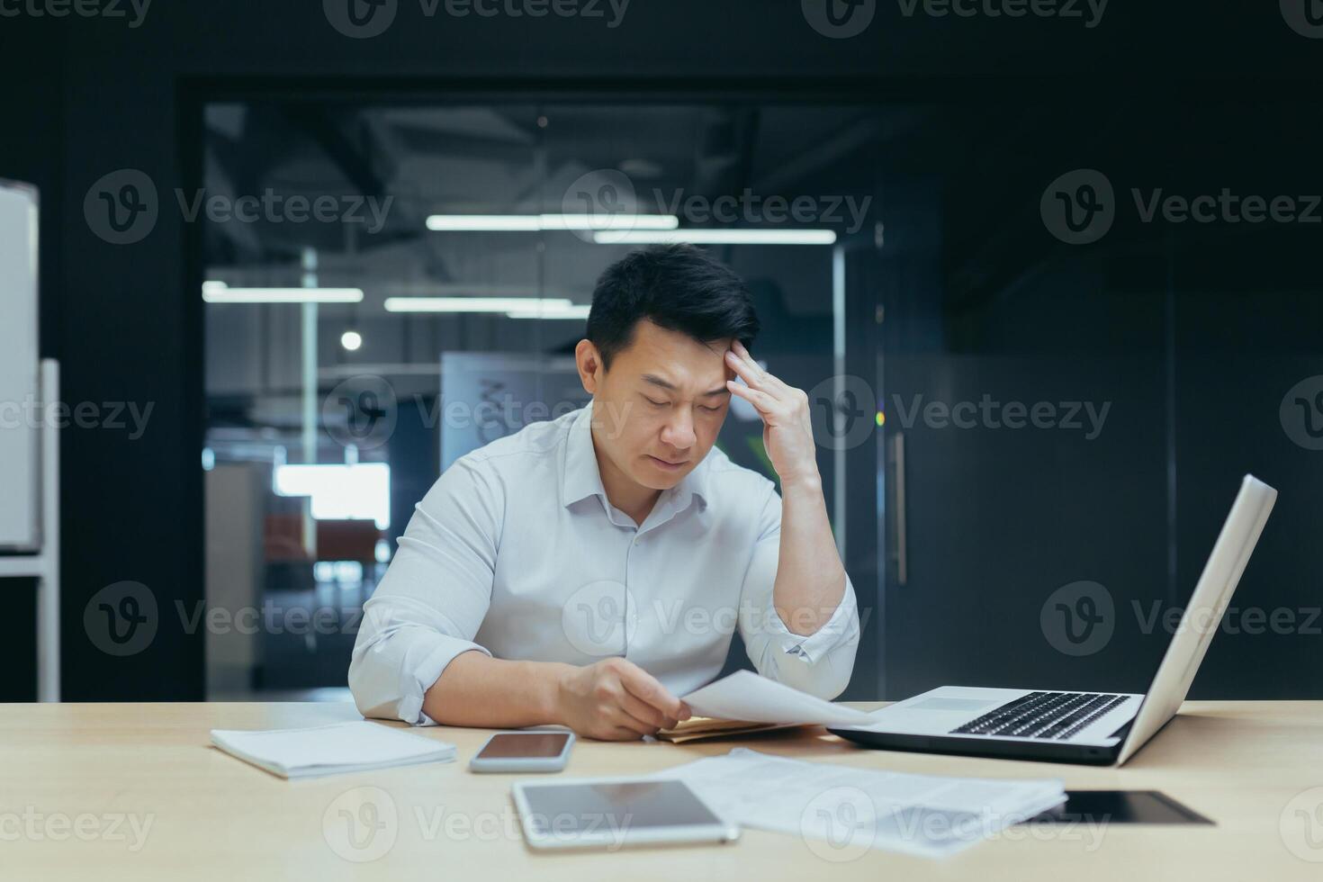 A hard day's work. A serious young Asian man sits tiredly in the office at the desk, works with documents, problems with accounts, holds his head with his hand. photo