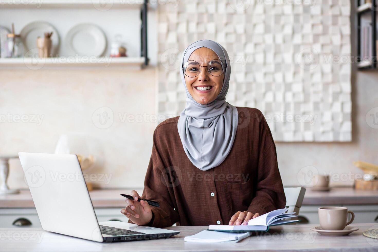 Portrait of a young Muslim female psychologist specialist in a hijab sitting at home in front of a laptop and making notes in a notebook. He consults and works online. Smiling looking at the camera. photo