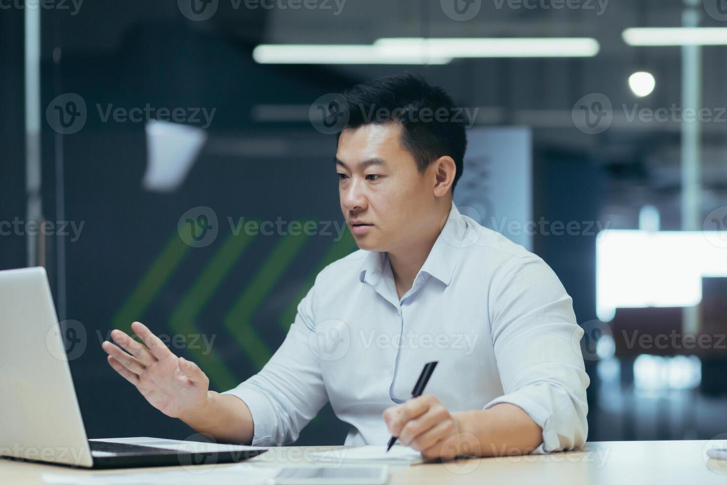 Worried Asian businessman sitting at the table in the office with laptop, talking on a call online, holding a pen, working with documents. solving the problem with the bank, bills, credit. photo