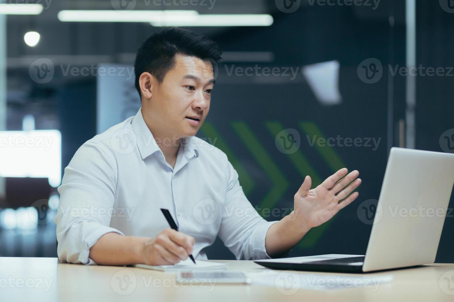 Asian businessman online meeting, looking at laptop screen and talking with colleagues, recording data in report, man call with paper work photo