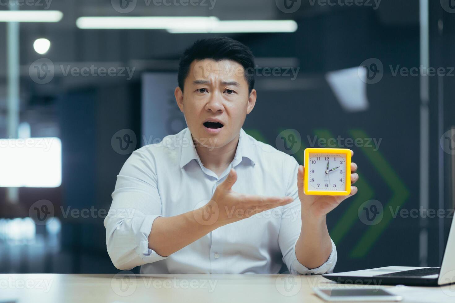 Time management. Young handsome asian businessman holding a desk clock in his hand, pointing to the clock, indicating the time, confused, late, looking at the camera photo