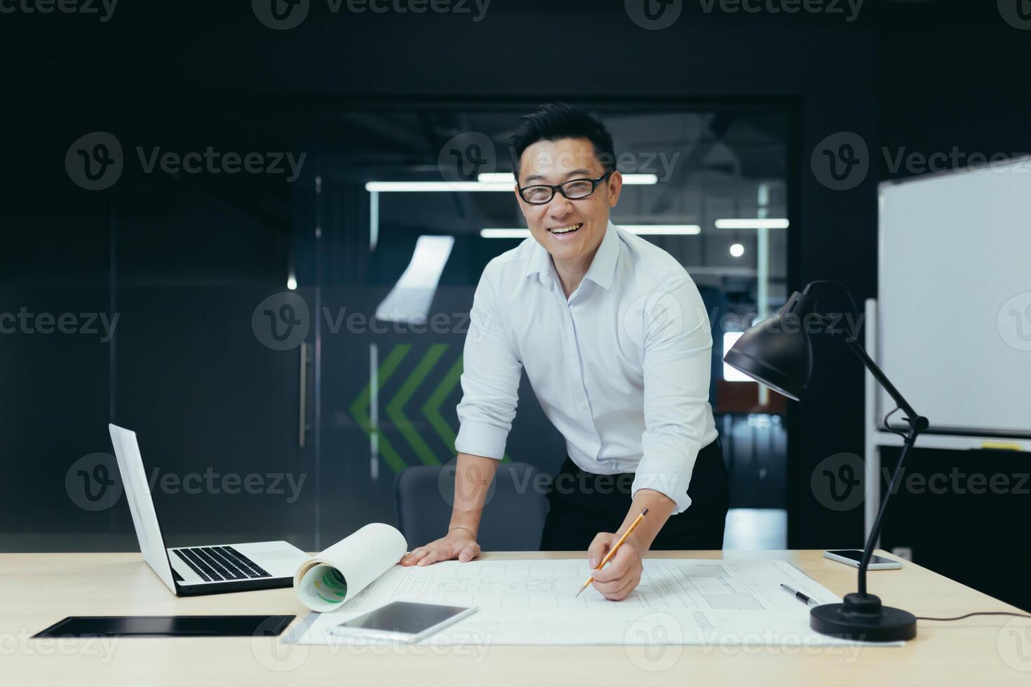 Portrait of successful and experienced Asian architect, man standing over table with plan drawing, smiling and looking at camera, working in modern design studio photo