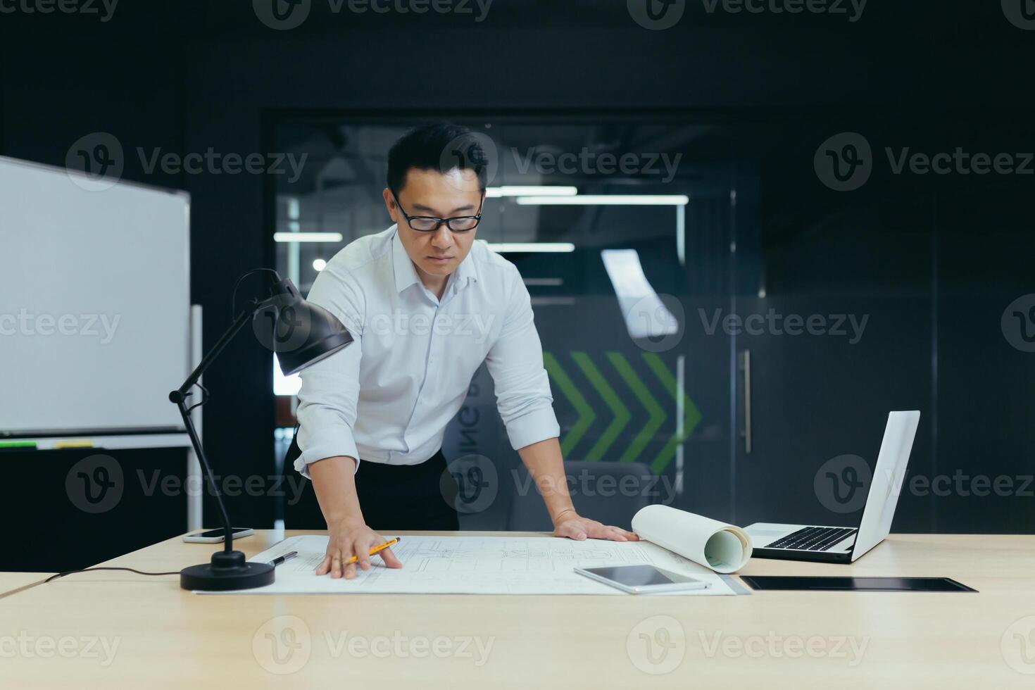 Handsome young man Asian architect, designer, engineer stands serious and focused in the office at the desk in a white shirt and glasses, works with plans, documents, drawings. photo