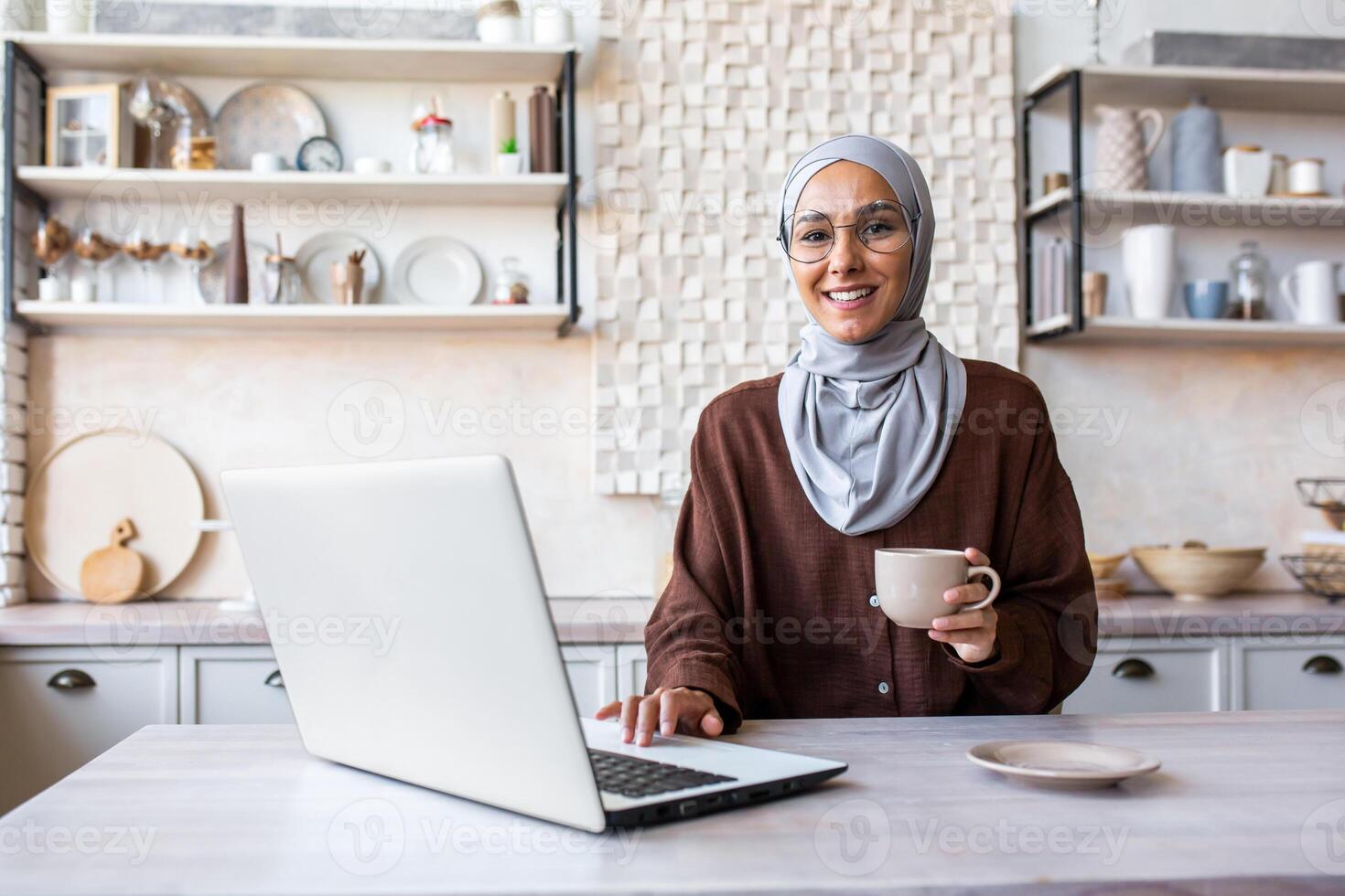 Muslim young woman in hijab working in kitchen at home at table. Drinks coffee, uses a laptop, takes a break, looks at the camera, smiles. photo