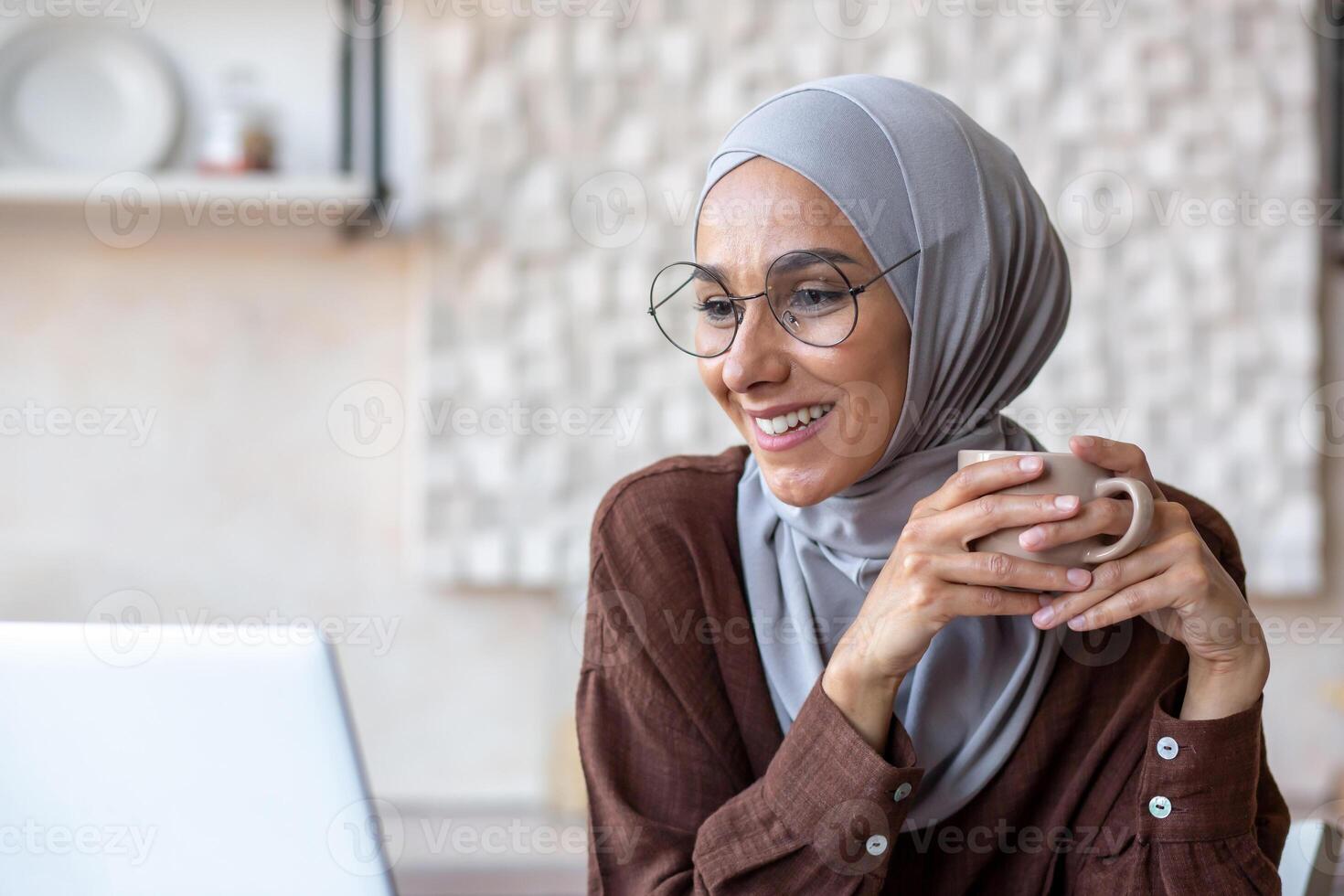 Close-up photo. Portrait of a young woman of oriental origin in a hijab sitting at home in the kitchen, smiling, looking at a laptop, holding a cup and drinking coffee. photo