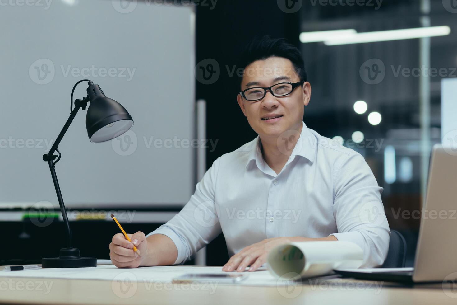 Portrait of a young handsome Asian man, architect, designer, freelancer. Sitting in the office at the table in glasses with a laptop. Makes a drawing, makes a plan. He looks at the camera, smiles. photo