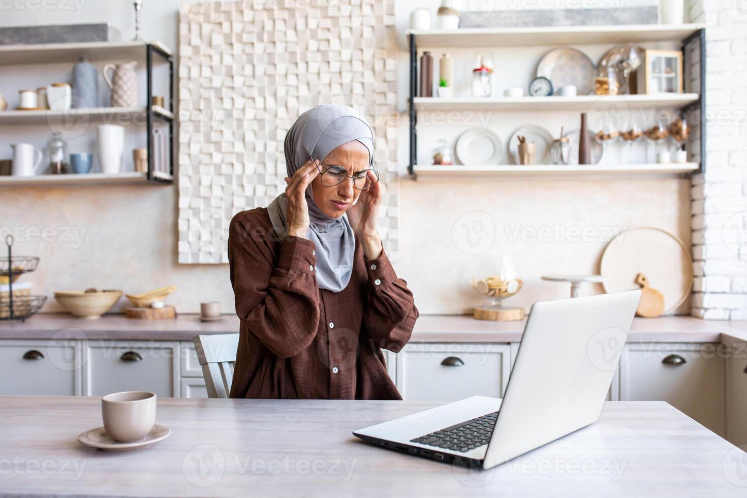 Tired young Muslim student in hijab studying remotely from home using laptop. He sits at the table and holds his head, feels severe pain, tension, and fatigue. Does a massage, rests. photo