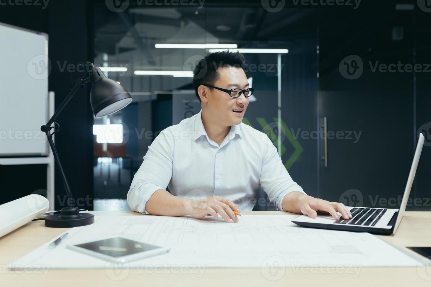 Smiling Asian male designer, engineer working in office at desk behind laptop and blueprints. photo
