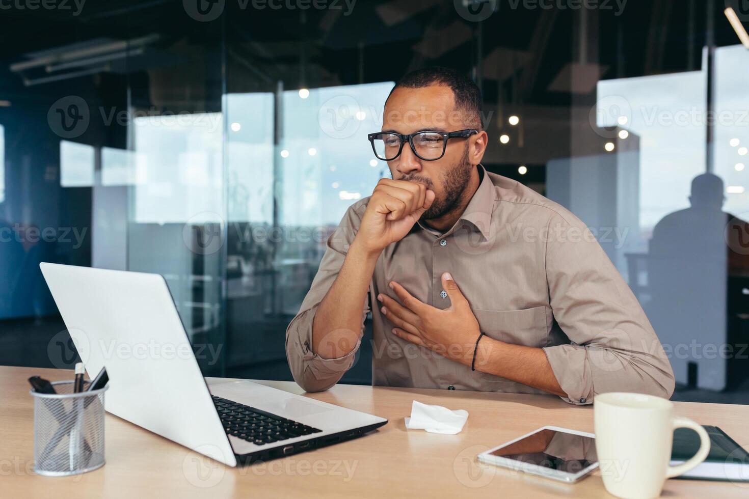 Businessman coughing at work in office, african american man in glasses and shirt sick at workplace using laptop at work. photo