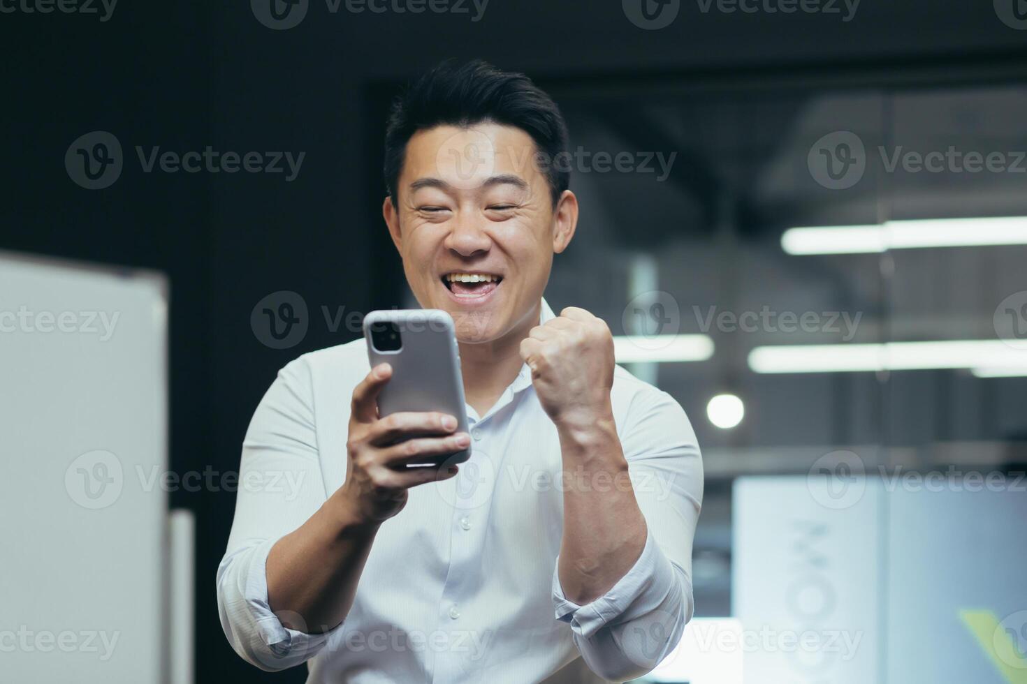 A break at work. A young Asian man holds a phone in his hands, plays games on the phone in the office at work, rejoices, shows with his hand yes,rejoices at the win, won a prize, rests, does not work. photo