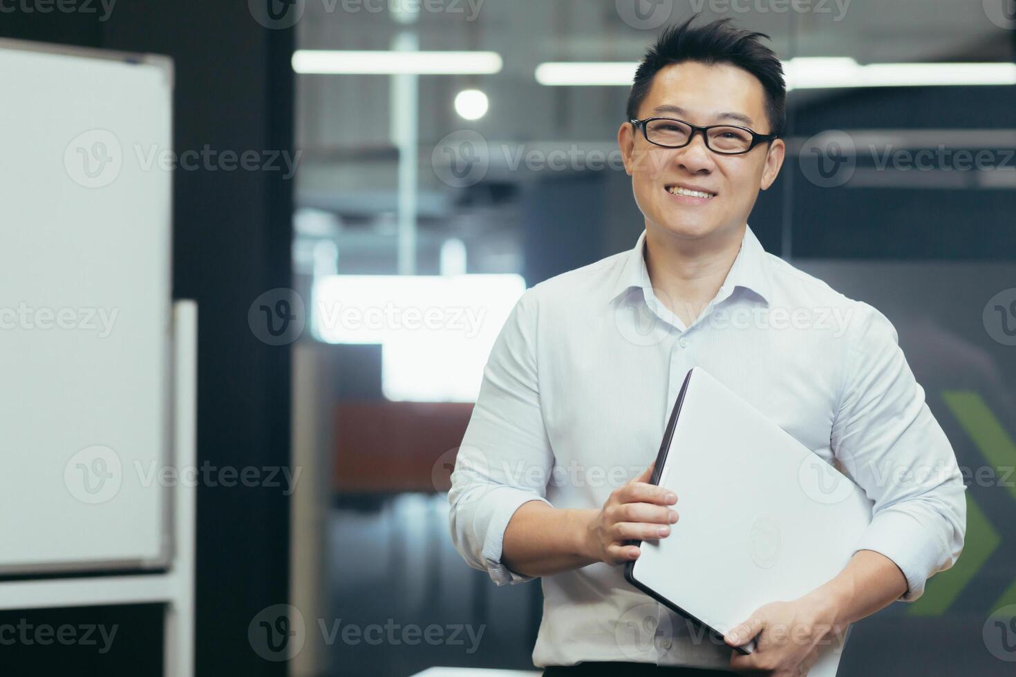 Portrait of a smiling handsome young Asian student. He is standing in the office of the campus wearing glasses and a white shirt, holding a folder with documents in his hands. photo