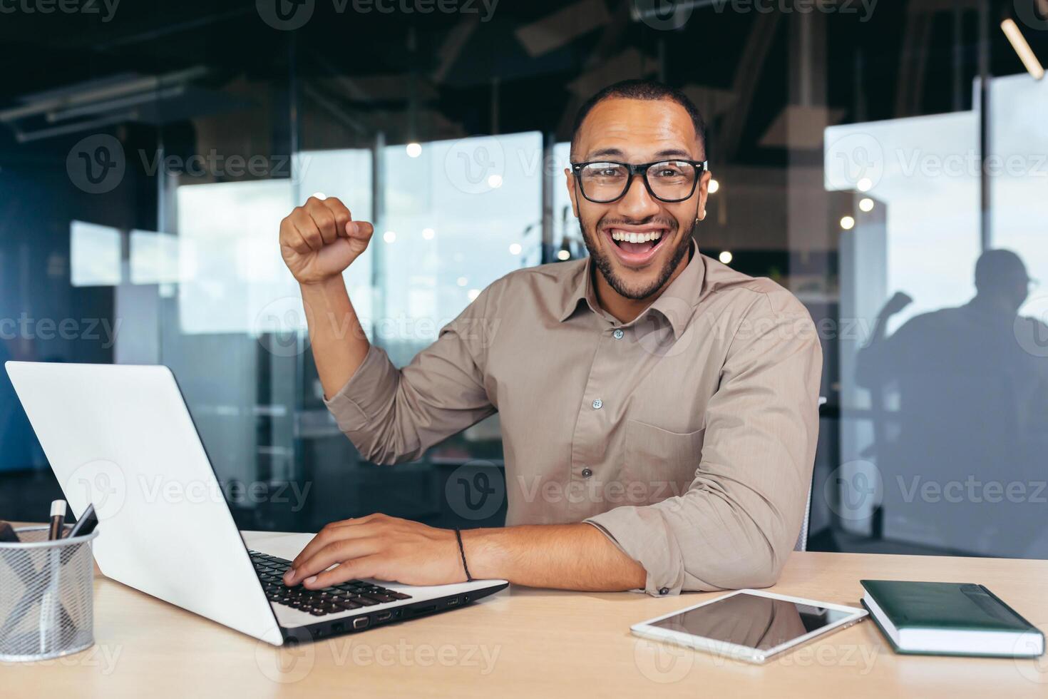 Portrait of successful hispanic executive, man in glasses smiling and looking at camera holding hand up gesture of achievement, good work result, victory and triumph. photo