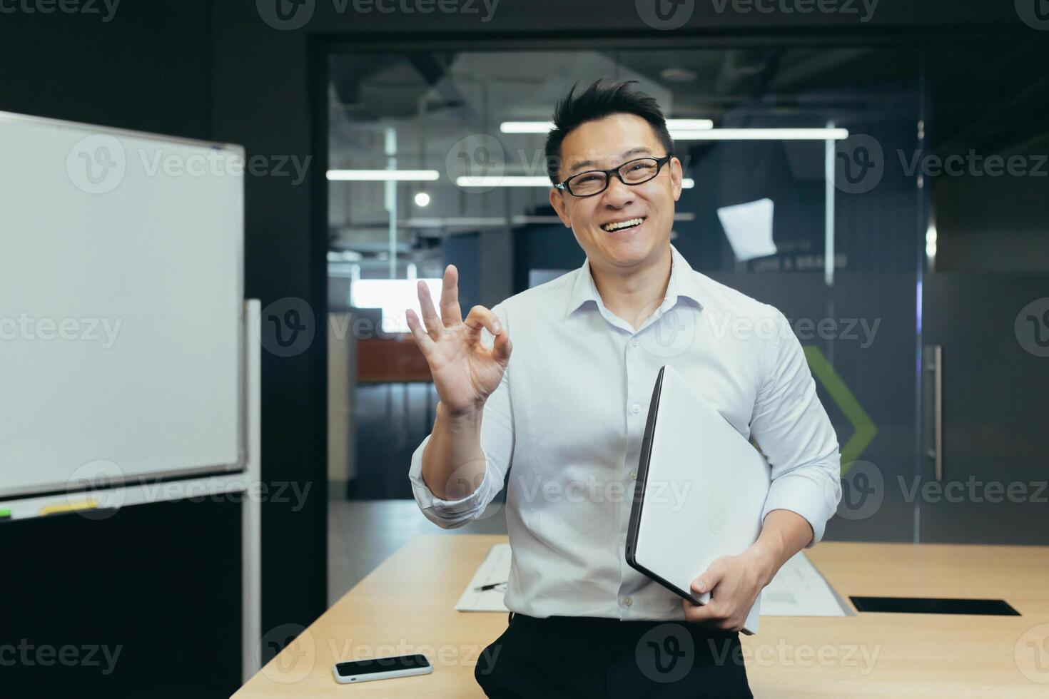 Portrait of a young handsome Asian student, intern, assistant. He stands with a folder in the office, wears glasses, smiles, points with his fingers photo