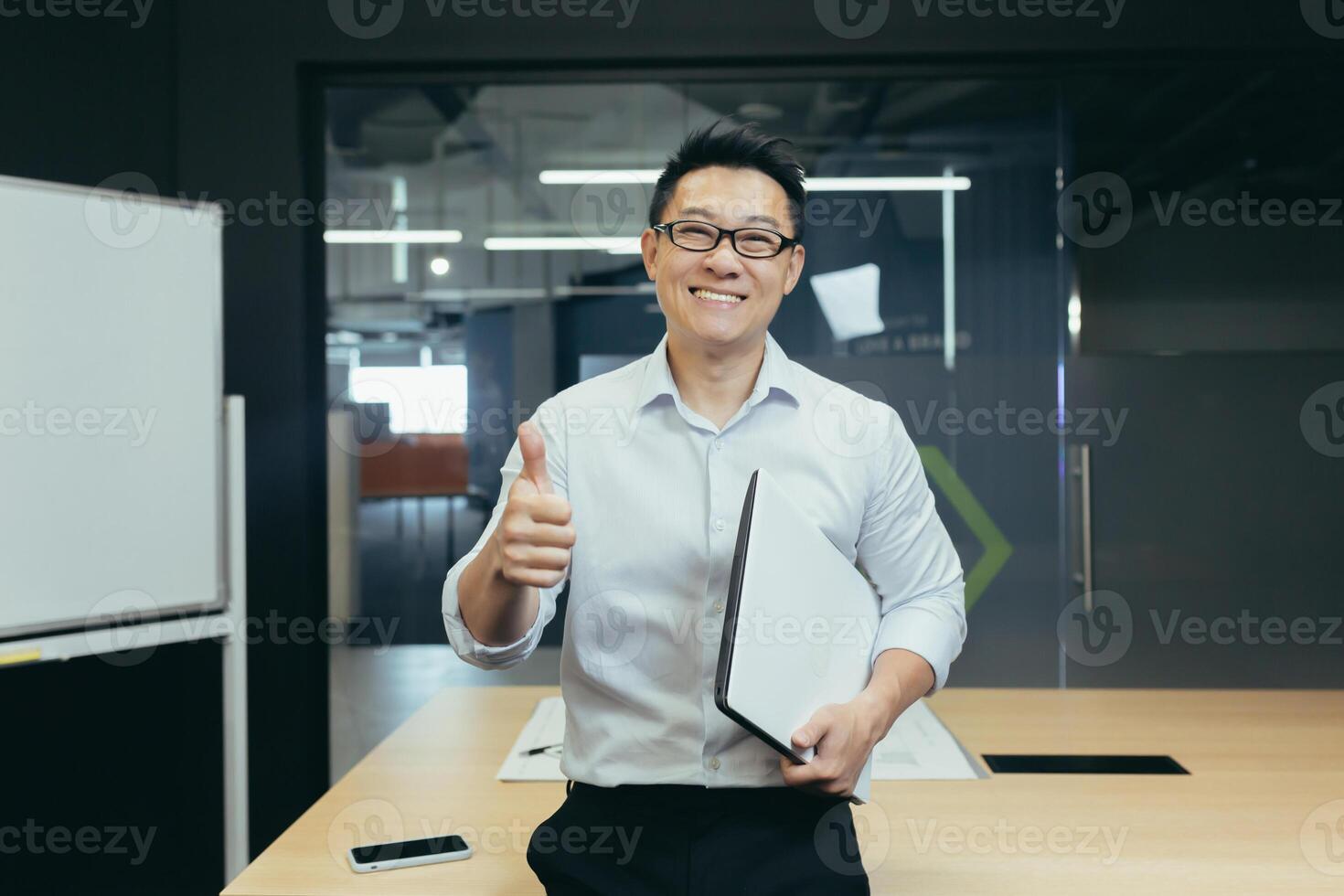 Portrait of successful asian teacher, man in modern classroom, looking at camera and smiling, wearing glasses holding laptop, showing thumbs up gesture affirmative photo