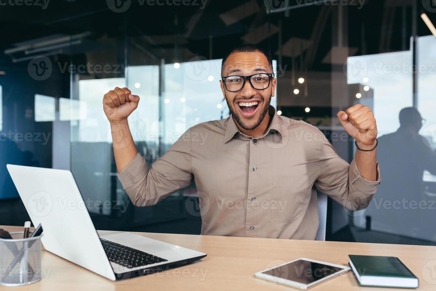 Happy and successful businessman celebrating achievement, portrait of man at work in office looking at camera and holding hands up, celebrating victory and triumph, employee using laptop. photo