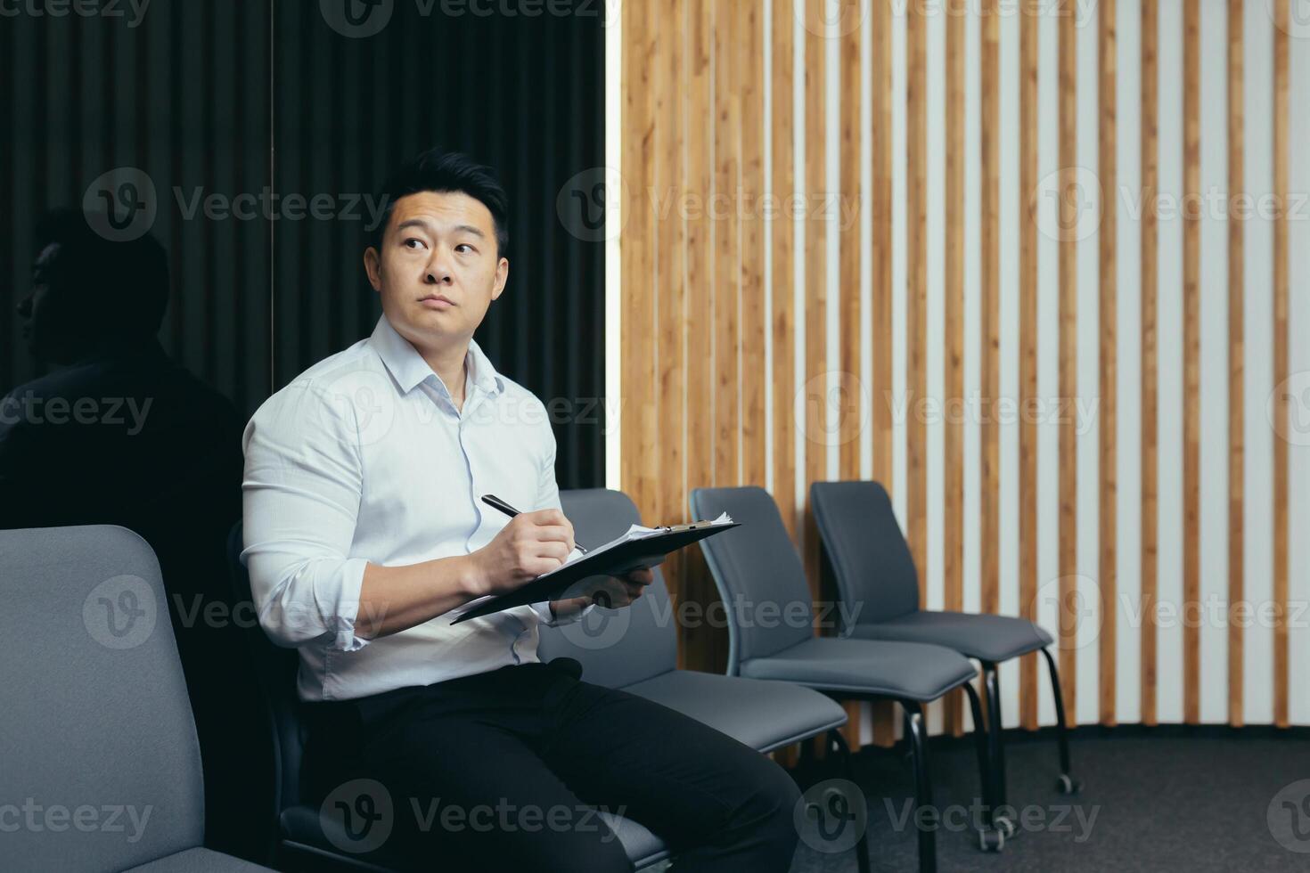 A serious young Asian man fills out a questionnaire for the reception desk, sits in the lobby of an office, hotel, hospital. He holds a folder with papers in his hands, looks to the side. photo