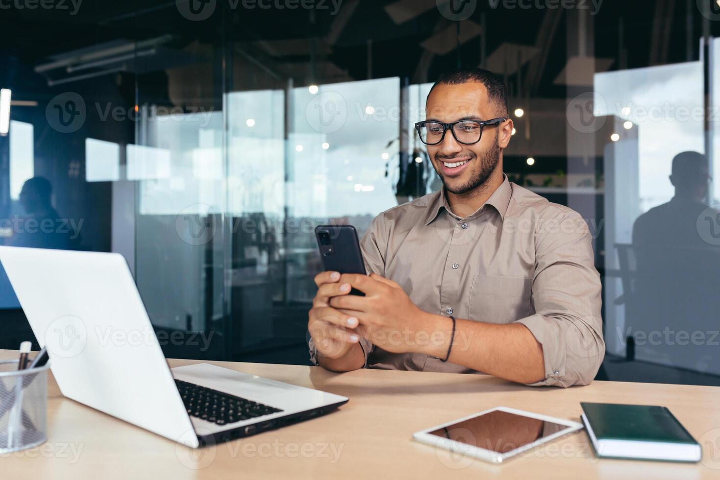 Happy and smiling businessman rejoicing at successful work results and achievement, man at work using phone reading online good news sitting at table inside office, using laptop at work. photo