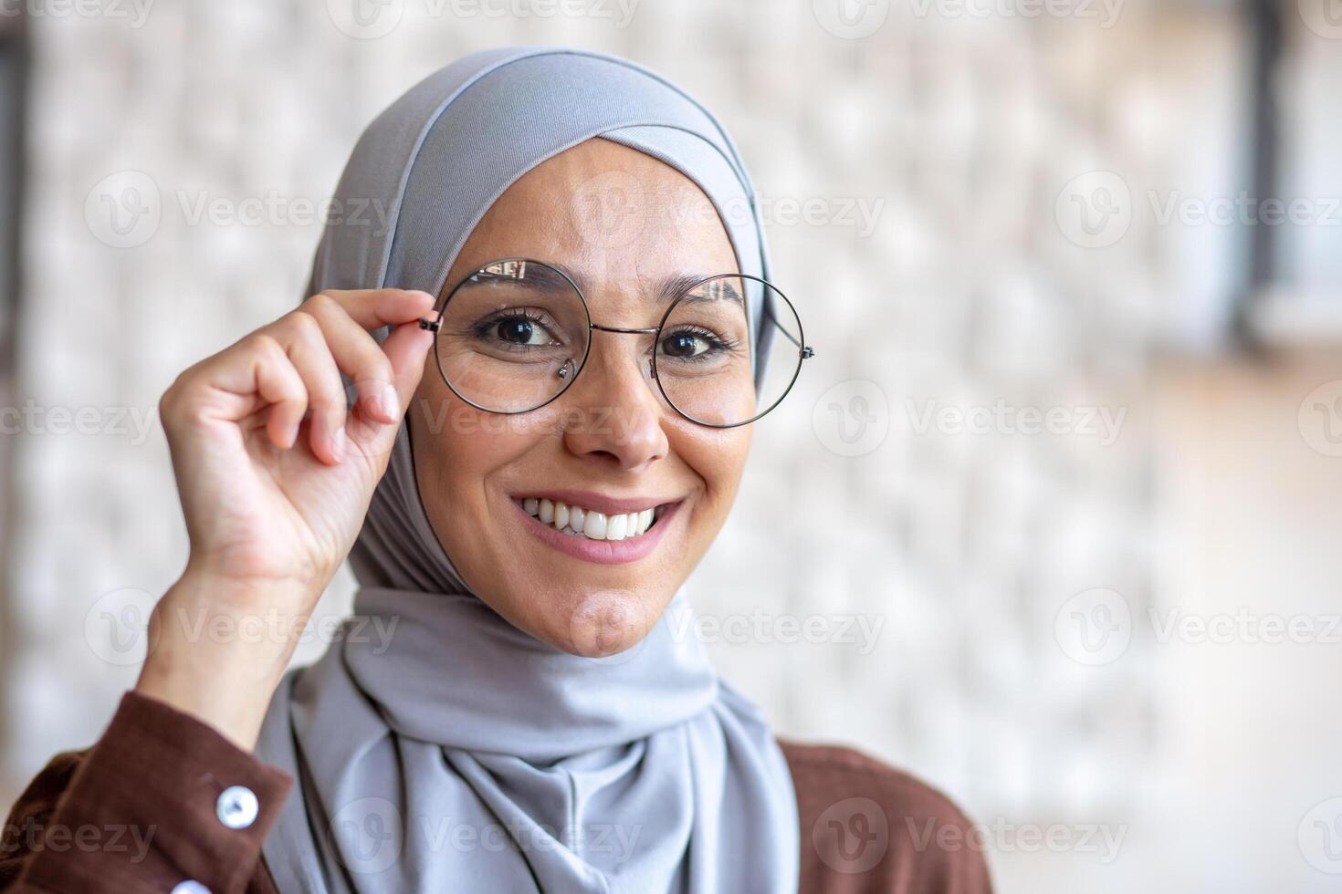 Close-up photo. Portrait of a young beautiful Muslim woman in a hijab smiling looking at the camera and adjusting her glasses with her hand. photo