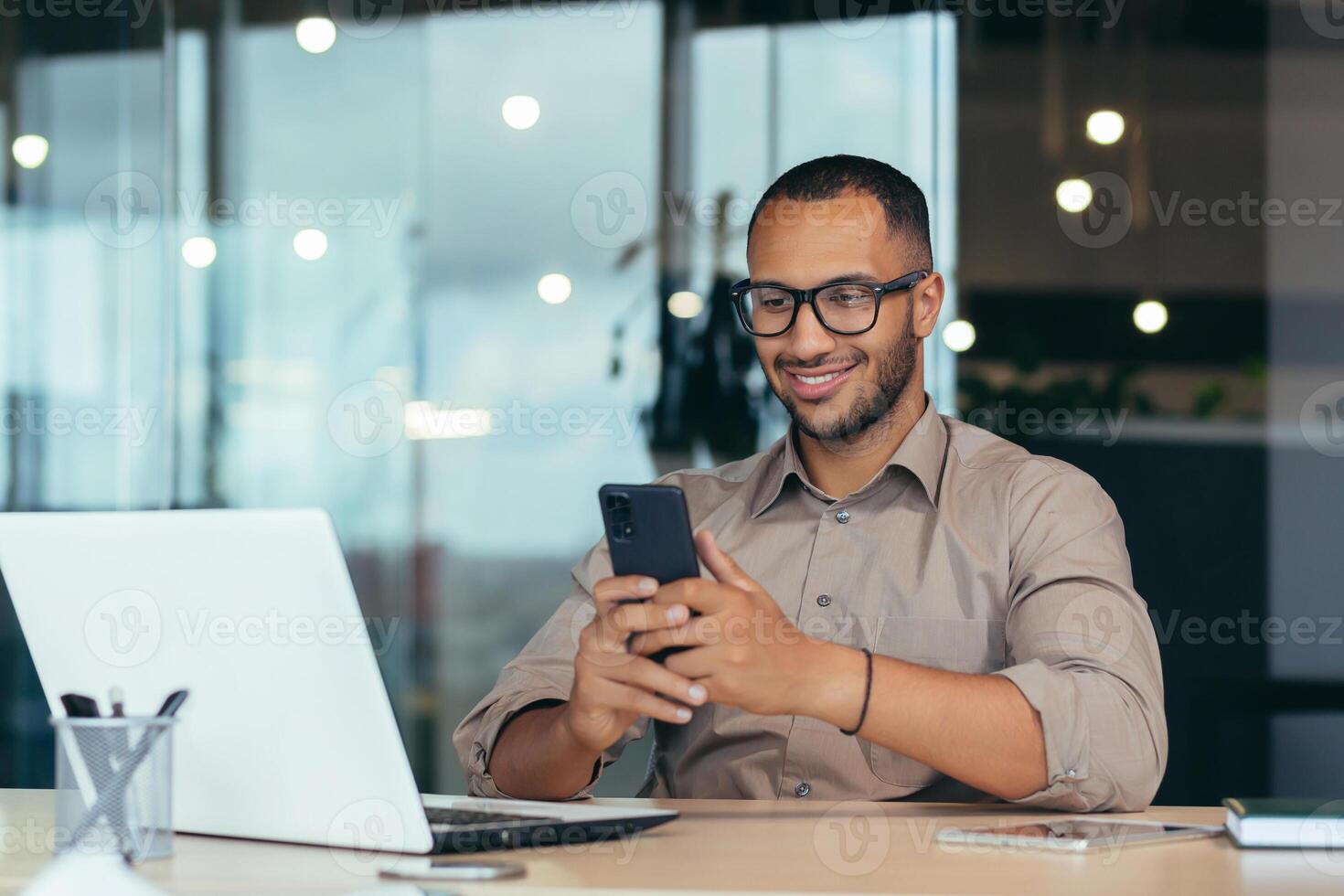 Happy african american businessman in glasses working inside modern office with laptop, man in shirt sitting at table browsing online pages using smartphone. photo