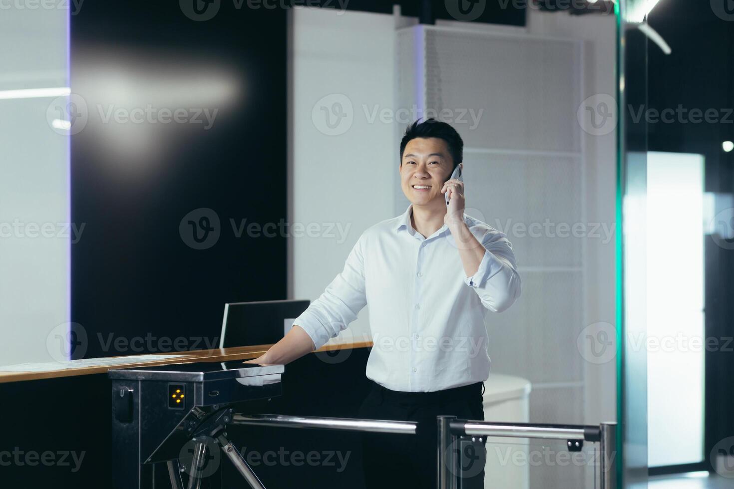 A young handsome Asian businessman passes through the security desk and pass in the morning to the office and business center for work. Talking on the phone, in a hurry, smiling photo
