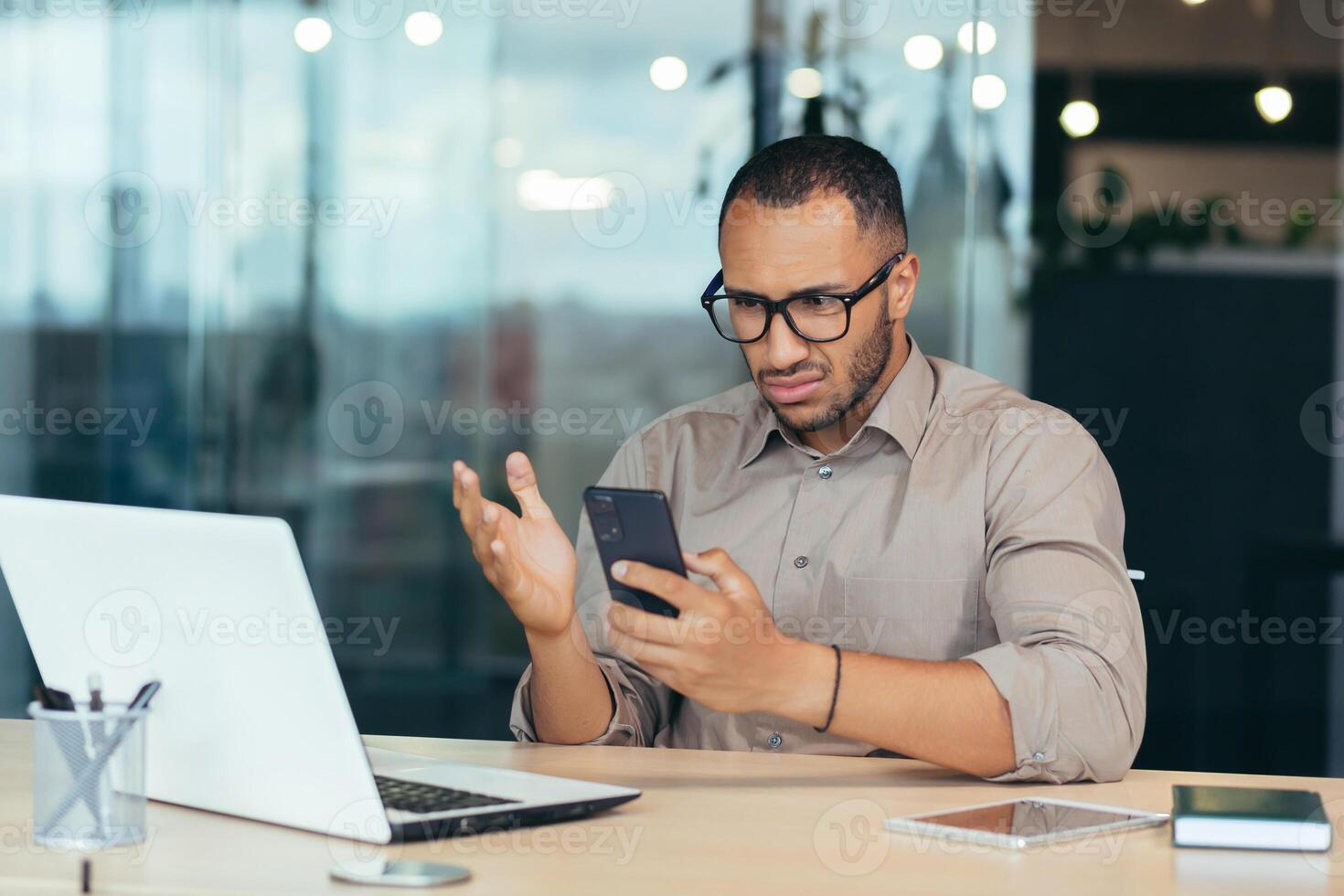 African american man received bad news notification online from phone, businessman in shirt working with laptop in middle of modern office building, man in glasses reading online letter. photo