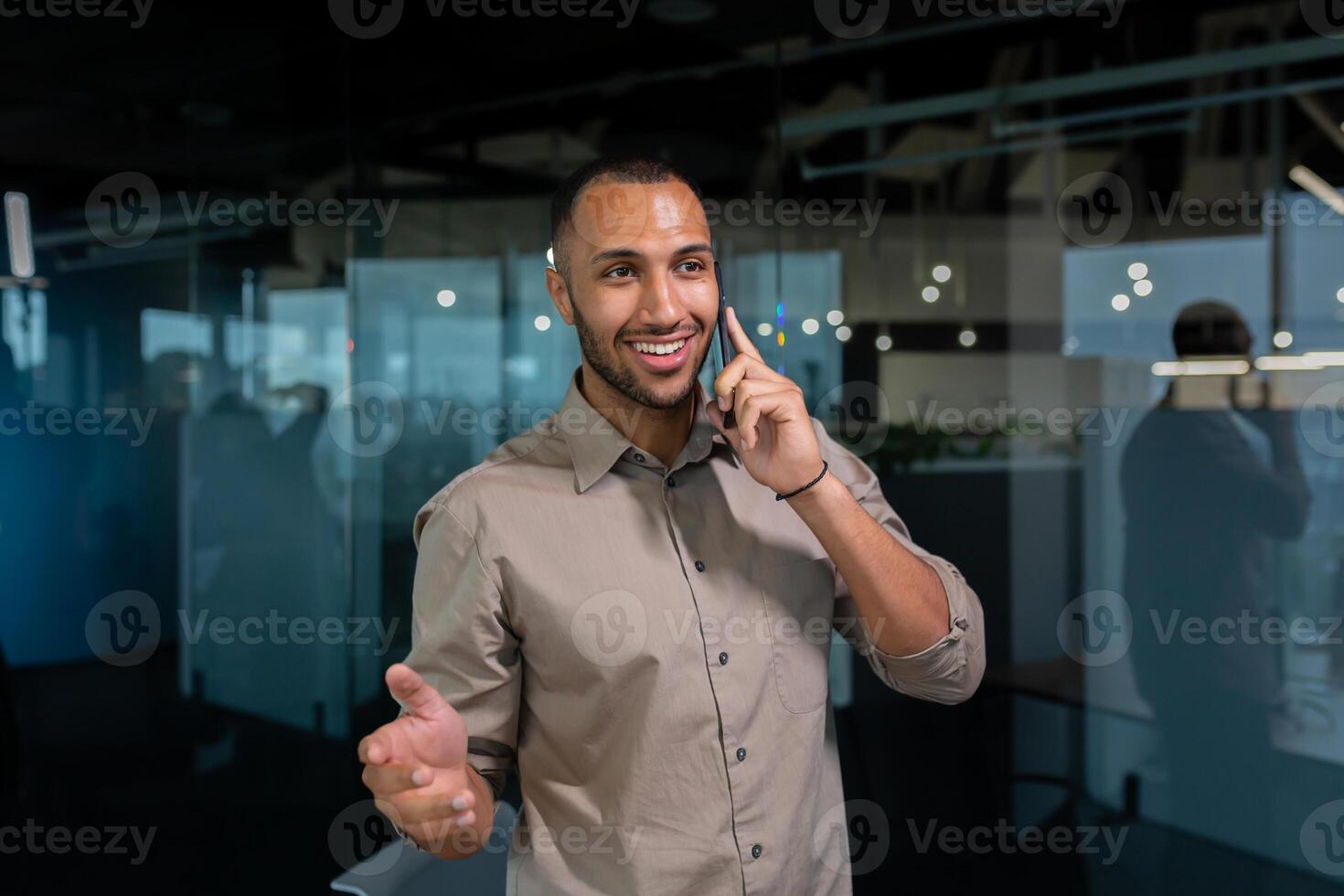 Cheerful and smiling businessman in a shirt talking on the phone inside the office, hispanic man standing near the window in the evening. photo