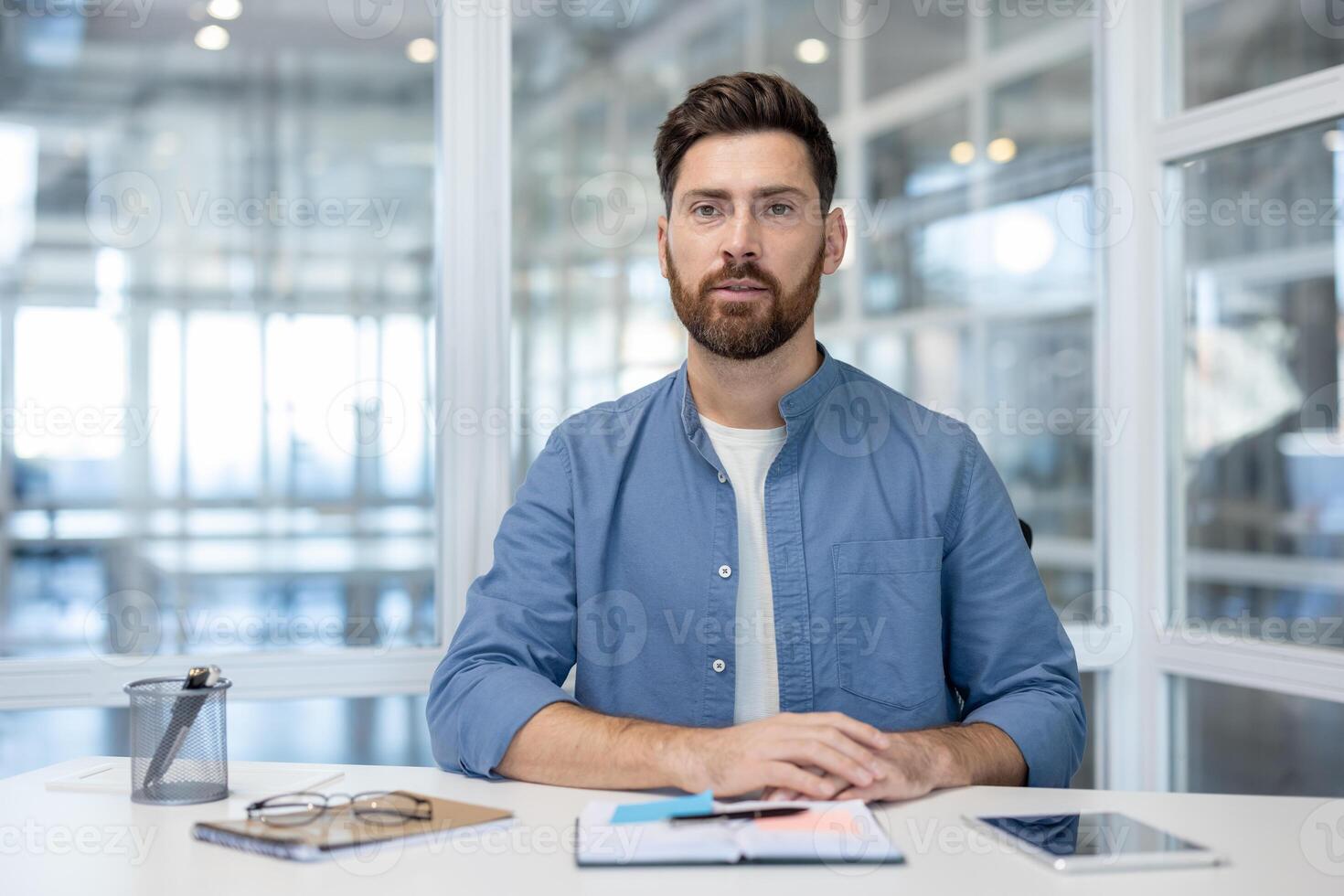 A well-groomed man with a beard sits at a desk, looking directly at the camera, possibly at a business meeting. photo