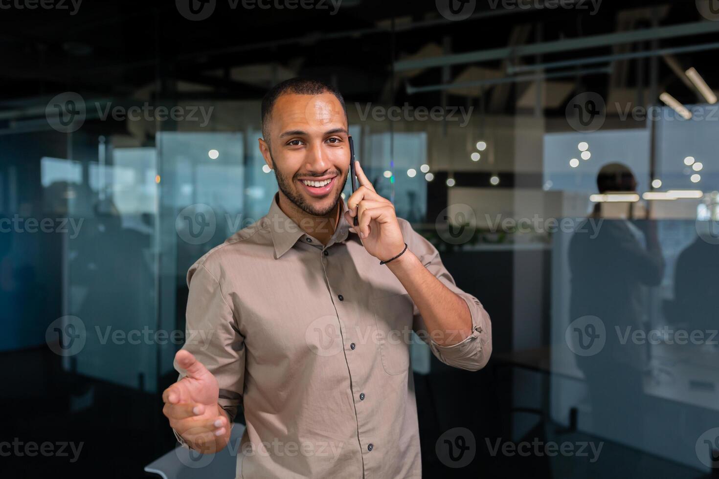 A young African American man is standing in the office and talking on the phone. gesturing with his hands, looking at the camera, smiling. photo