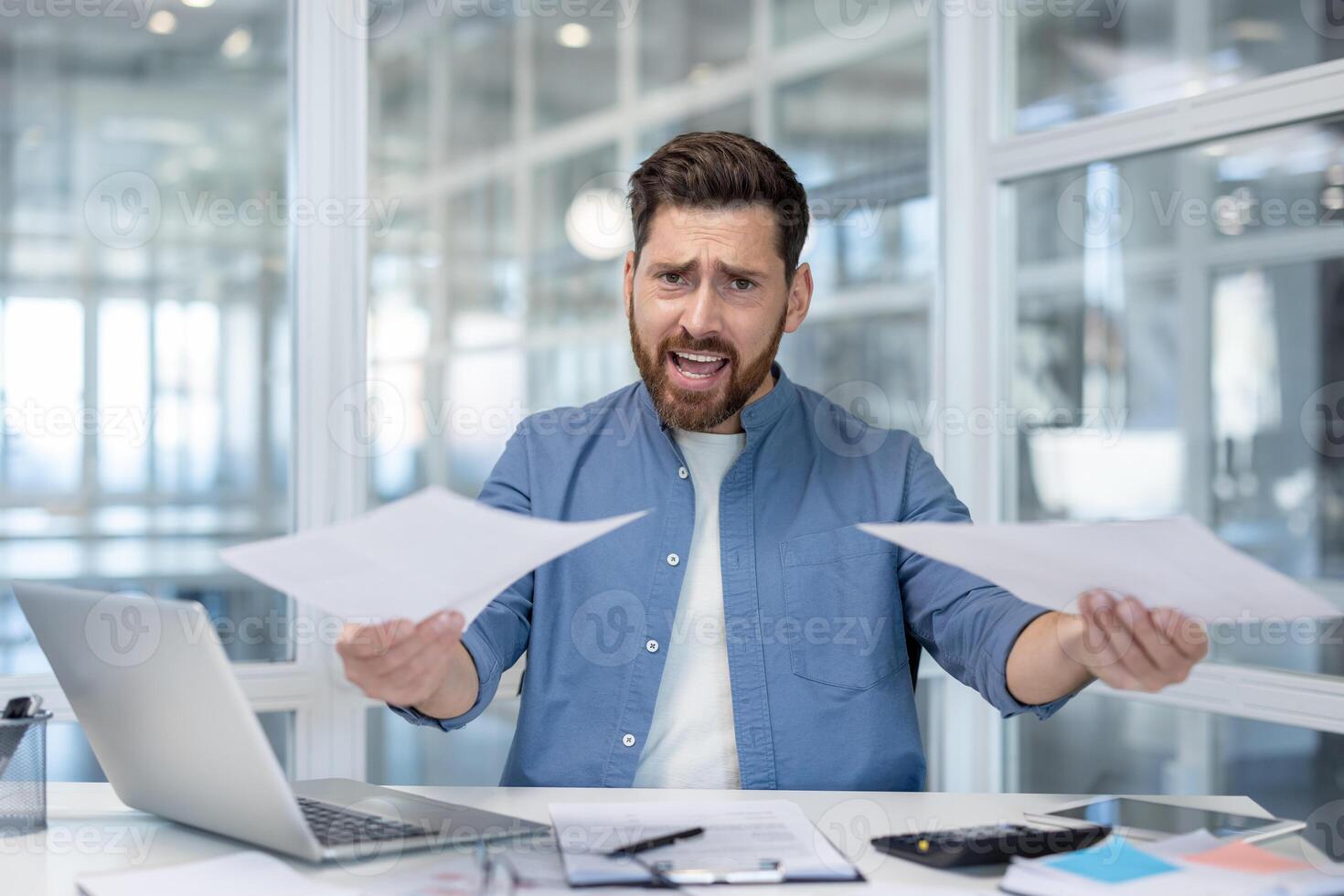 A frustrated businessman holds documents, looking stressed and overwhelmed at his office desk, surrounded by paperwork and technology. photo