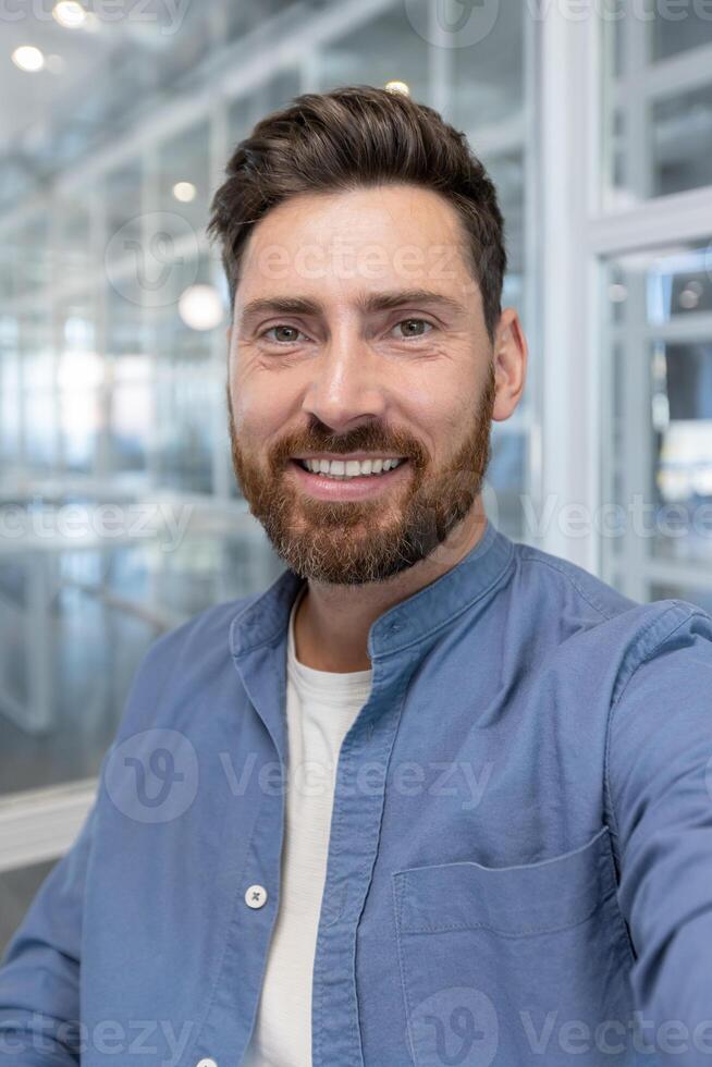 A smiling, bearded man in a blue shirt poses for a headshot indoors, showcasing his friendly and approachable demeanor. photo