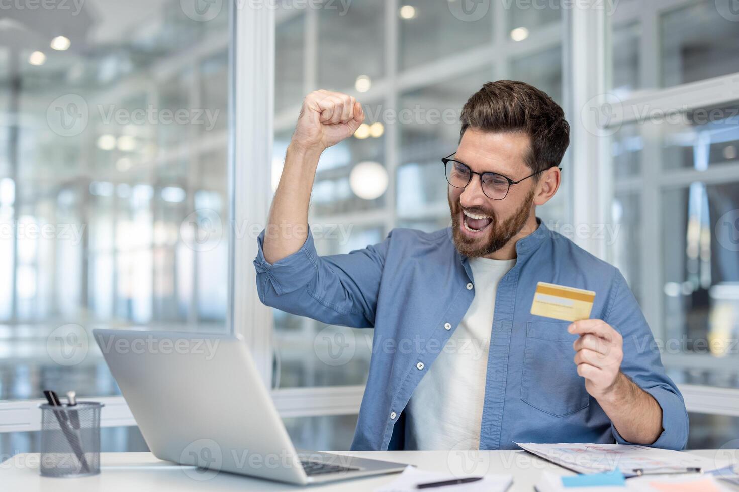 A man celebrates online payment success with a card in hand, sitting at a desk in front of a laptop, happy. photo