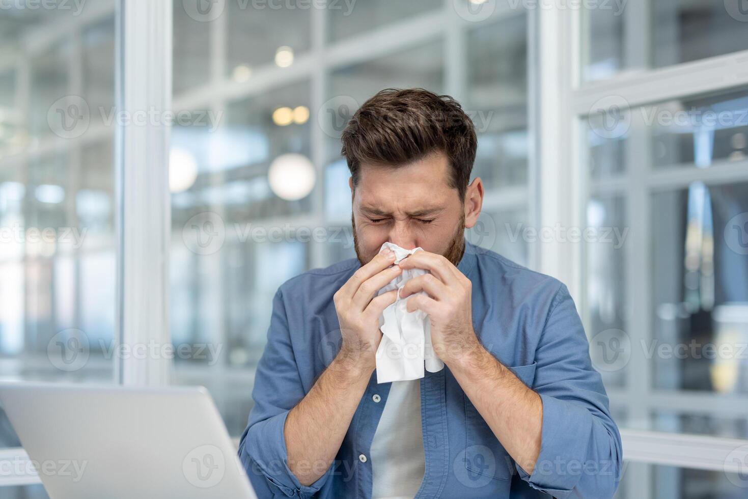 A man blowing his nose at work with a tissue, showing cold or allergy symptoms. Laptop on the desk visible. photo