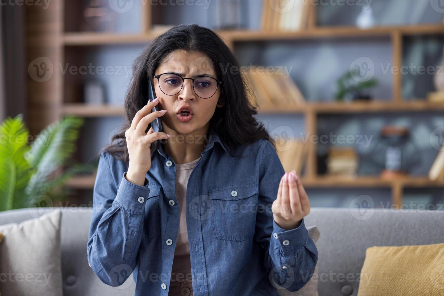 An angry young woman wearing glasses vehemently argues on her smartphone while gesturing expressively at home. The background is blurred. photo