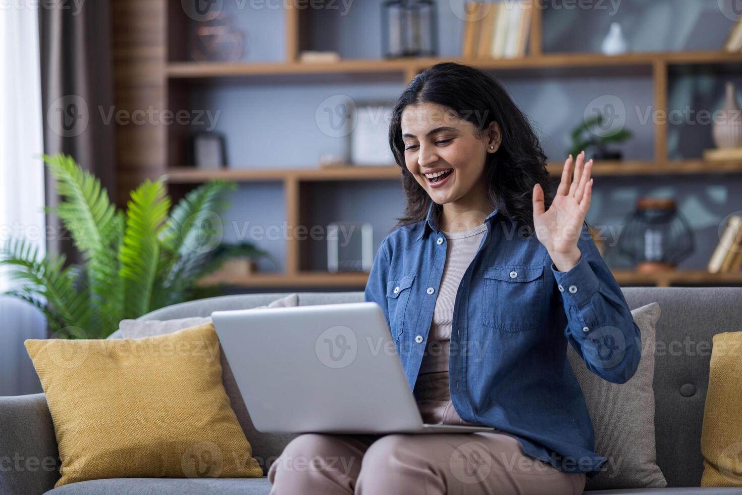 Young Indian girl sitting on the couch at home, holding a laptop on her lap, talking on a call, greeting and waving at the camera. photo