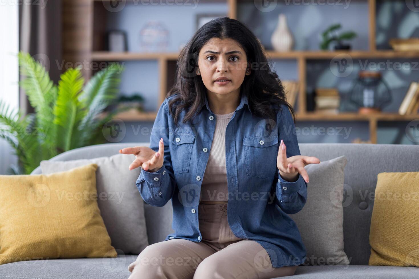 Upset Indian woman sitting on a sofa with a concerned expression. She is gesturing with her hands while looking directly at the camera. photo