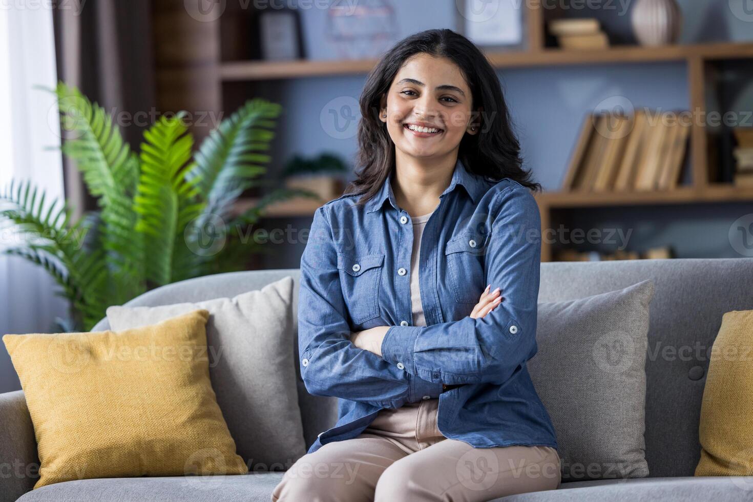 A beautiful brunette woman with arms crossed sitting on a couch in her living room with a fern and bookshelf in the background. photo