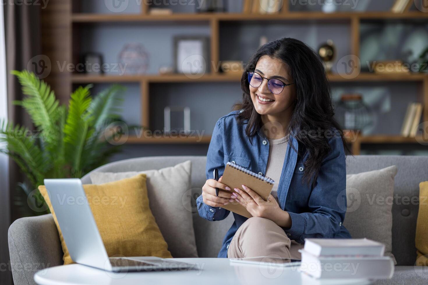 A young Muslim woman sits on the couch at home in front of a laptop, taking notes in a notebook, studying remotely. photo
