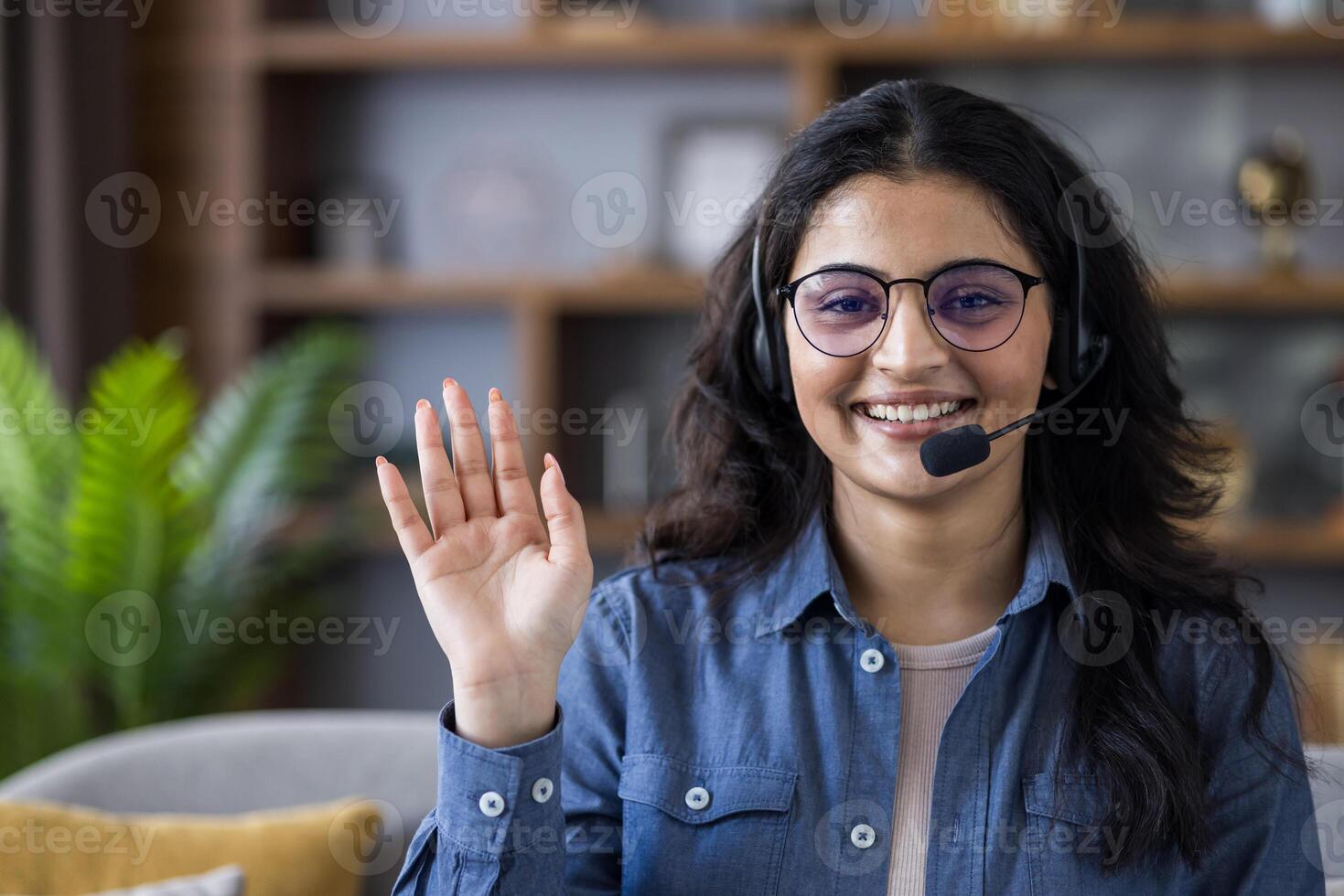 Friendly young woman wearing a headset smiles and waves during a virtual call, creating a warm and professional connection from home. photo