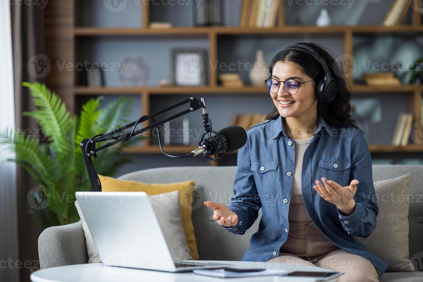 Smiling young Muslim woman in headset sitting on sofa at home in front of microphone and talking on laptop camera online. photo