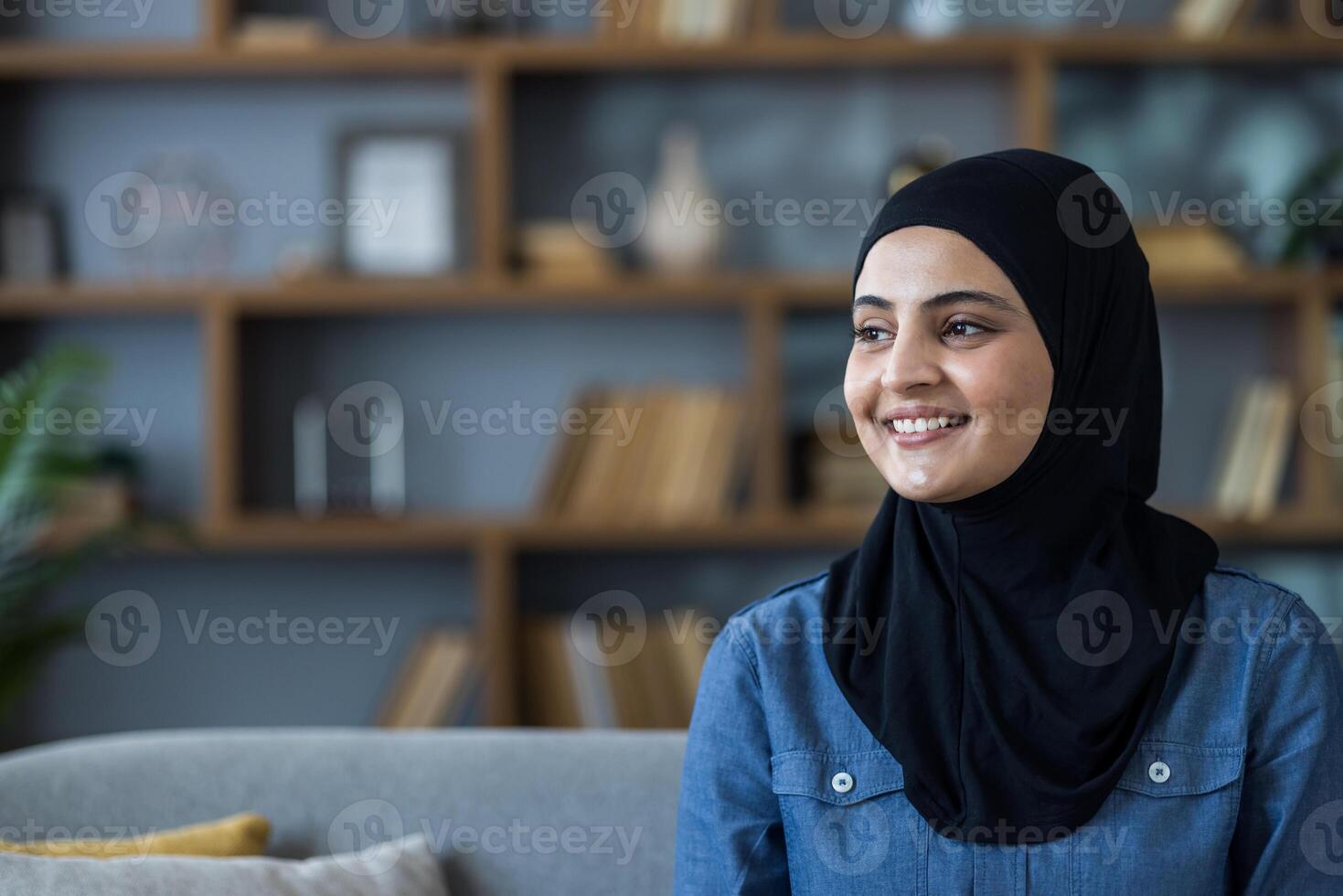 Close-up photo of a young Muslim woman wearing a black hijab sitting on a sofa at home and looking, to the side with a smile.