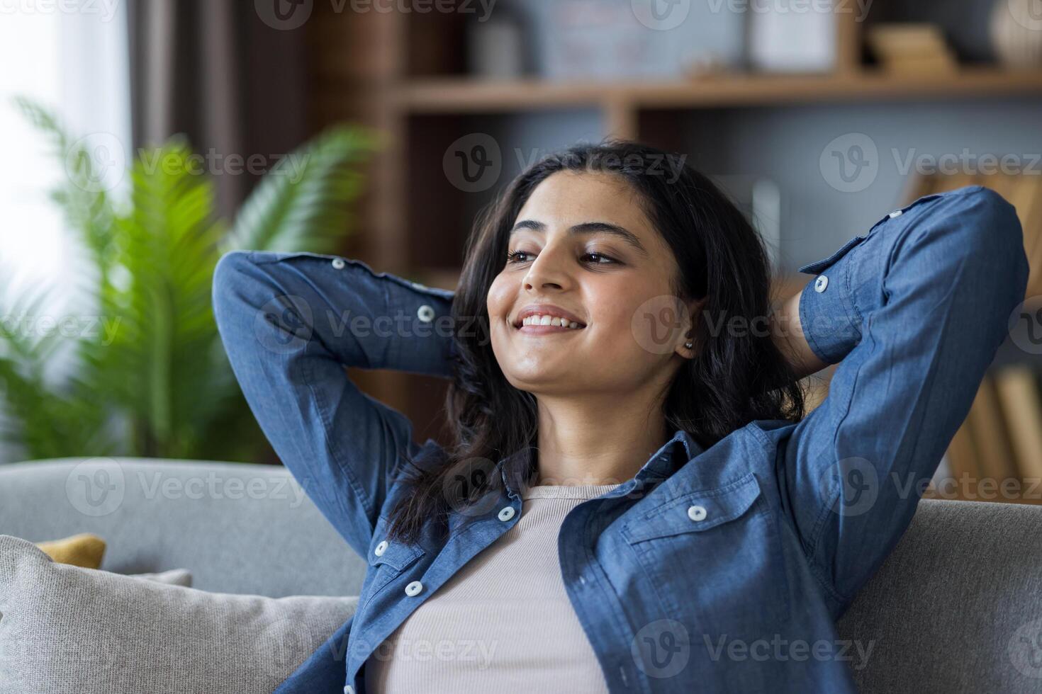 A close-up photo of a young Indian woman sitting on a couch, arms behind her head, leaning to the side, smiling and resting, satisfied with the result.