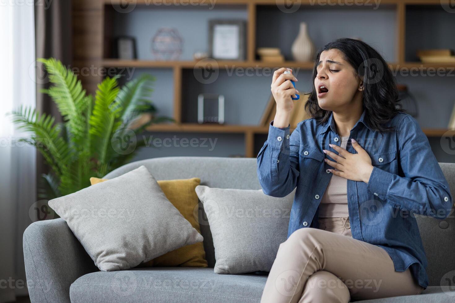 Young woman using an inhaler while sitting on a couch in her living room, experiencing an asthma attack and shortness of breath. photo