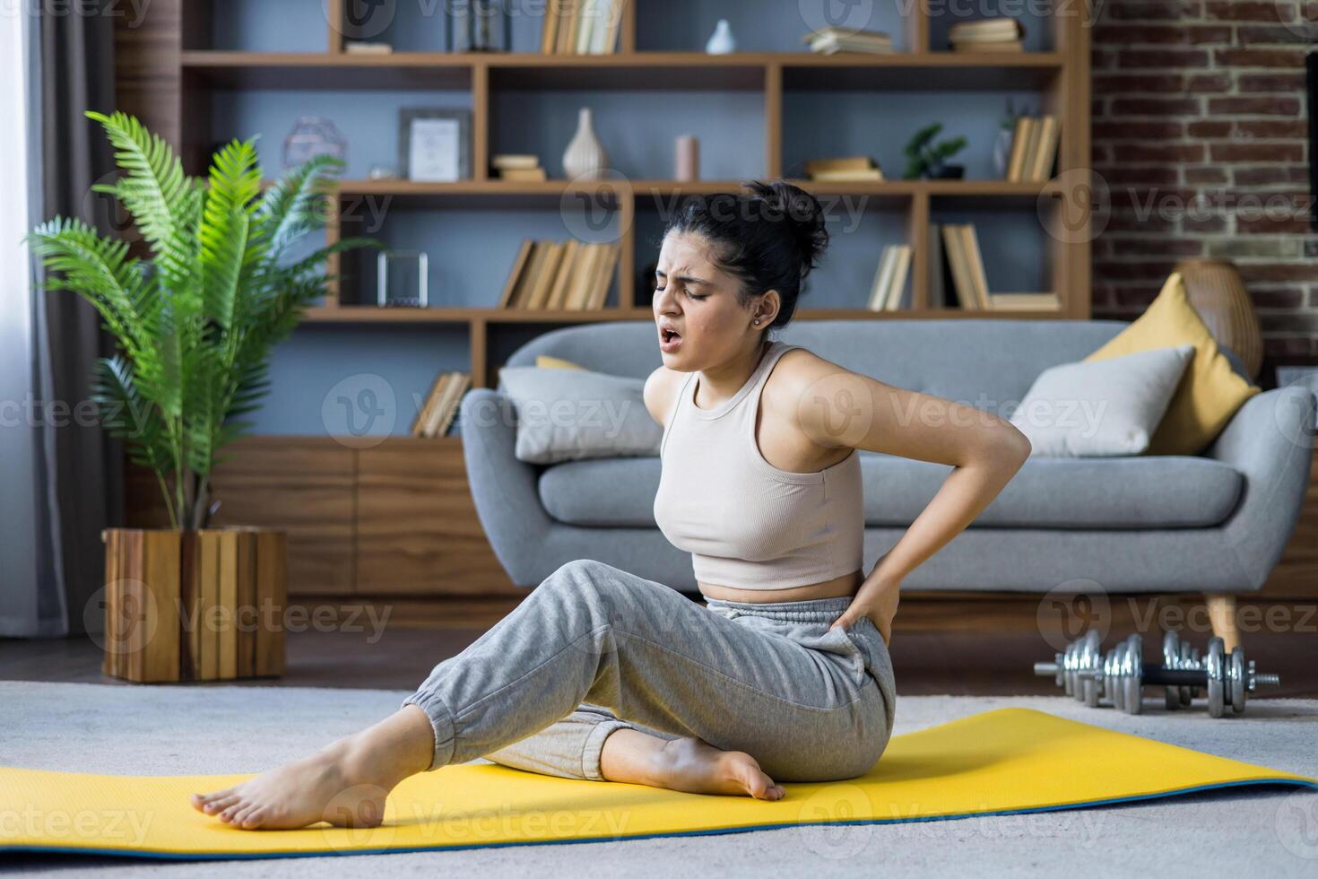 A young woman experiences back discomfort after a workout at home, sitting on a yoga mat, holding her back in pain, and wincing. photo