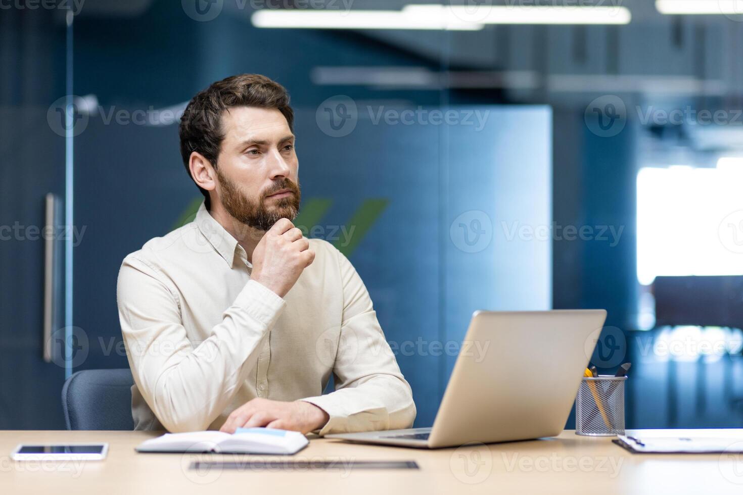 Photo of a serious young man sitting in the office at the workplace with a laptop and thoughtfully looking ahead, holding his hand by his beard.