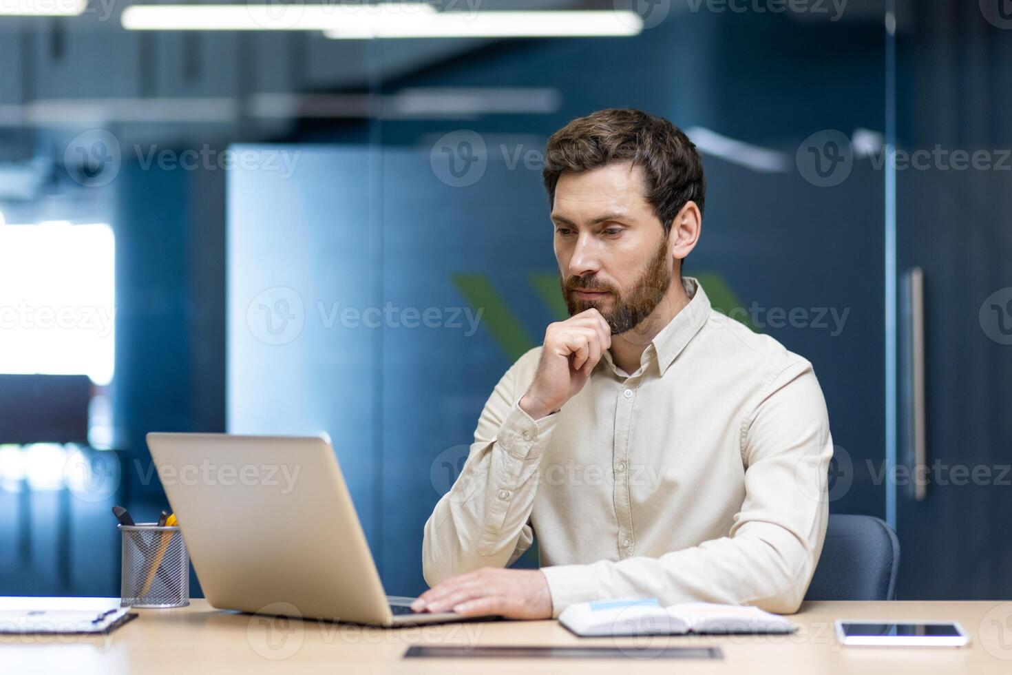 A young serious business man is sitting in the office at a desk and is concentrating on working on a laptop. photo