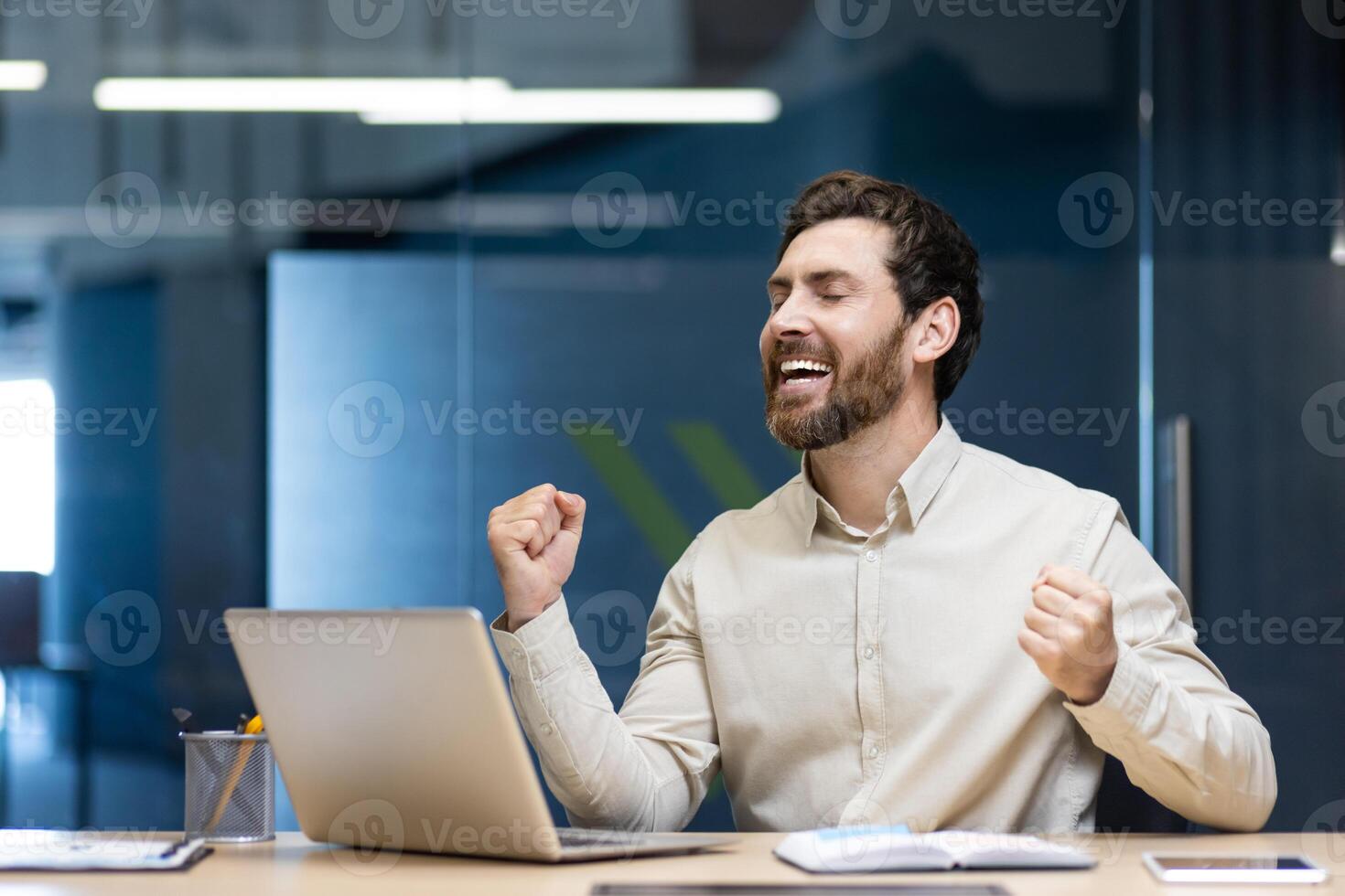 A happy young man businessman sits in the office at the table and rejoices at the success and result, raising his hands up. photo