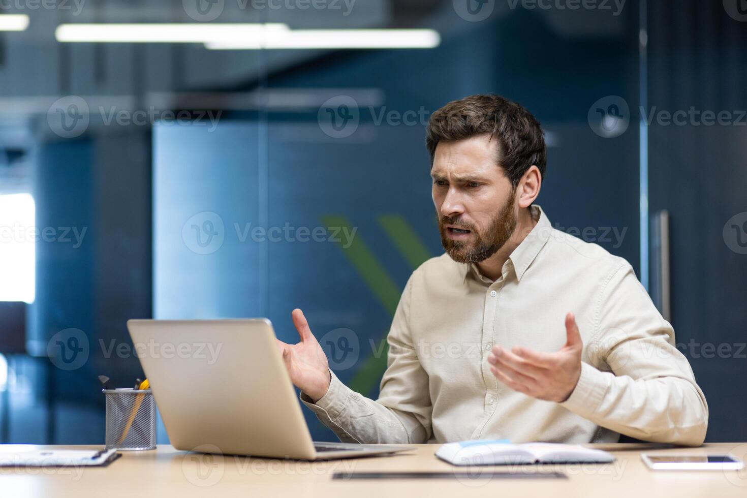 Frustrated and sad young businessman man sitting at desk in office and talking on call on laptop gesturing with hands. photo