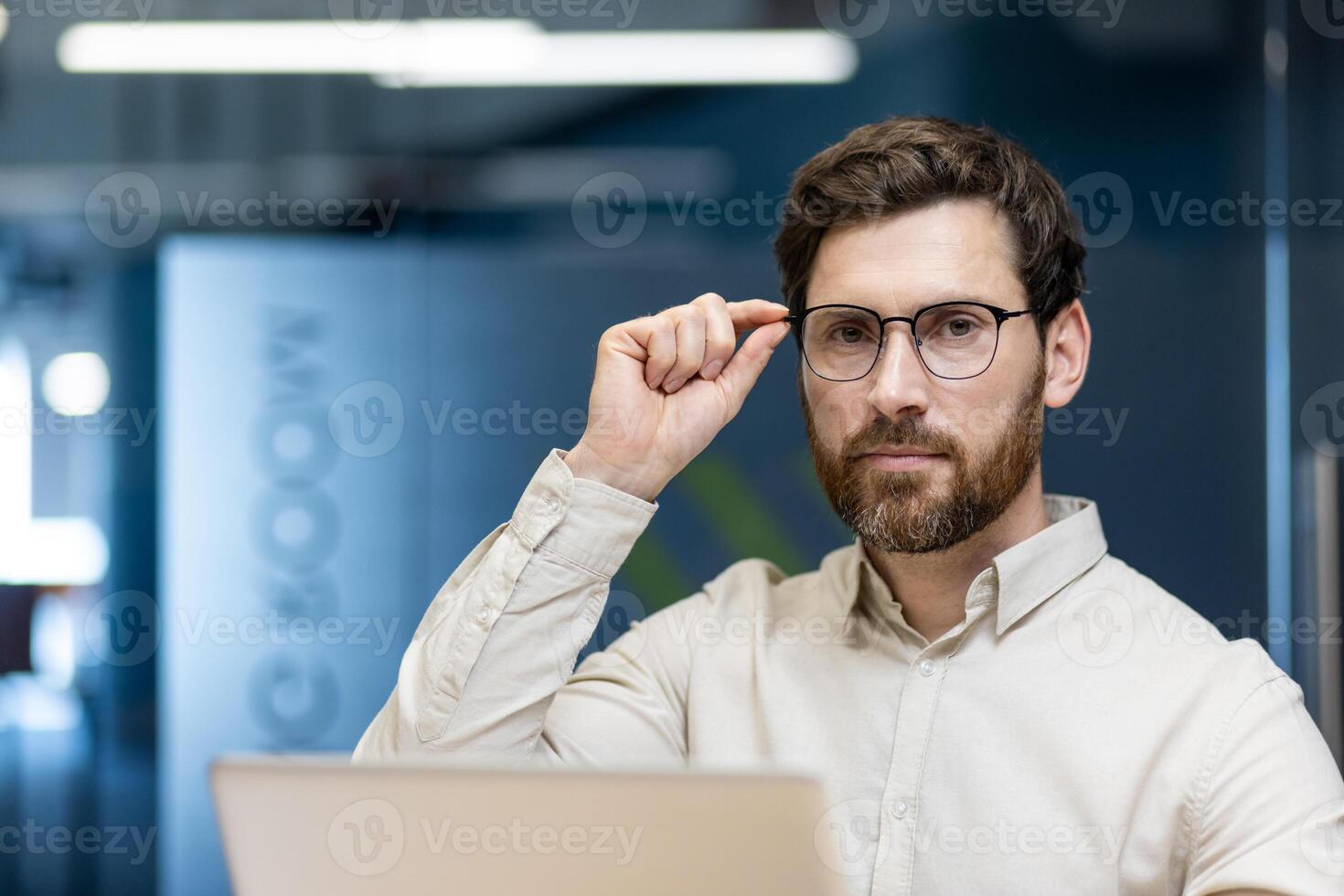 Close-up portrait of a serious young man sitting in the office at the workplace, working on a laptop, looking at the camera, holding glasses with his hand. photo