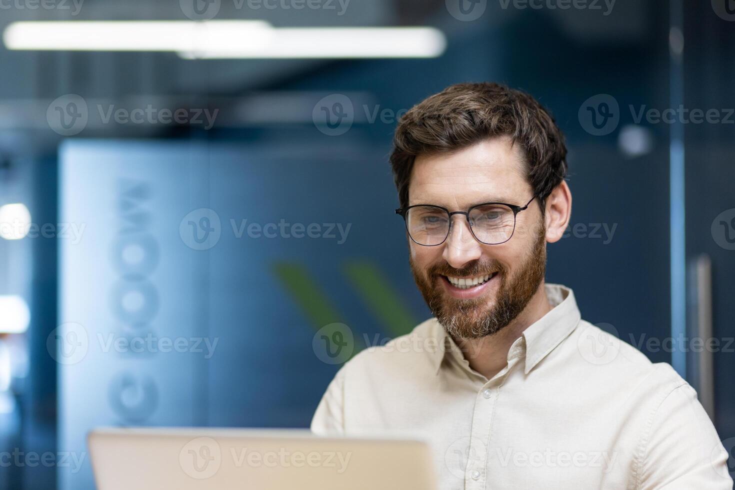 Close-up photo of a young smiling man in glasses and a light shirt sitting in the office at the workplace and working on a laptop.