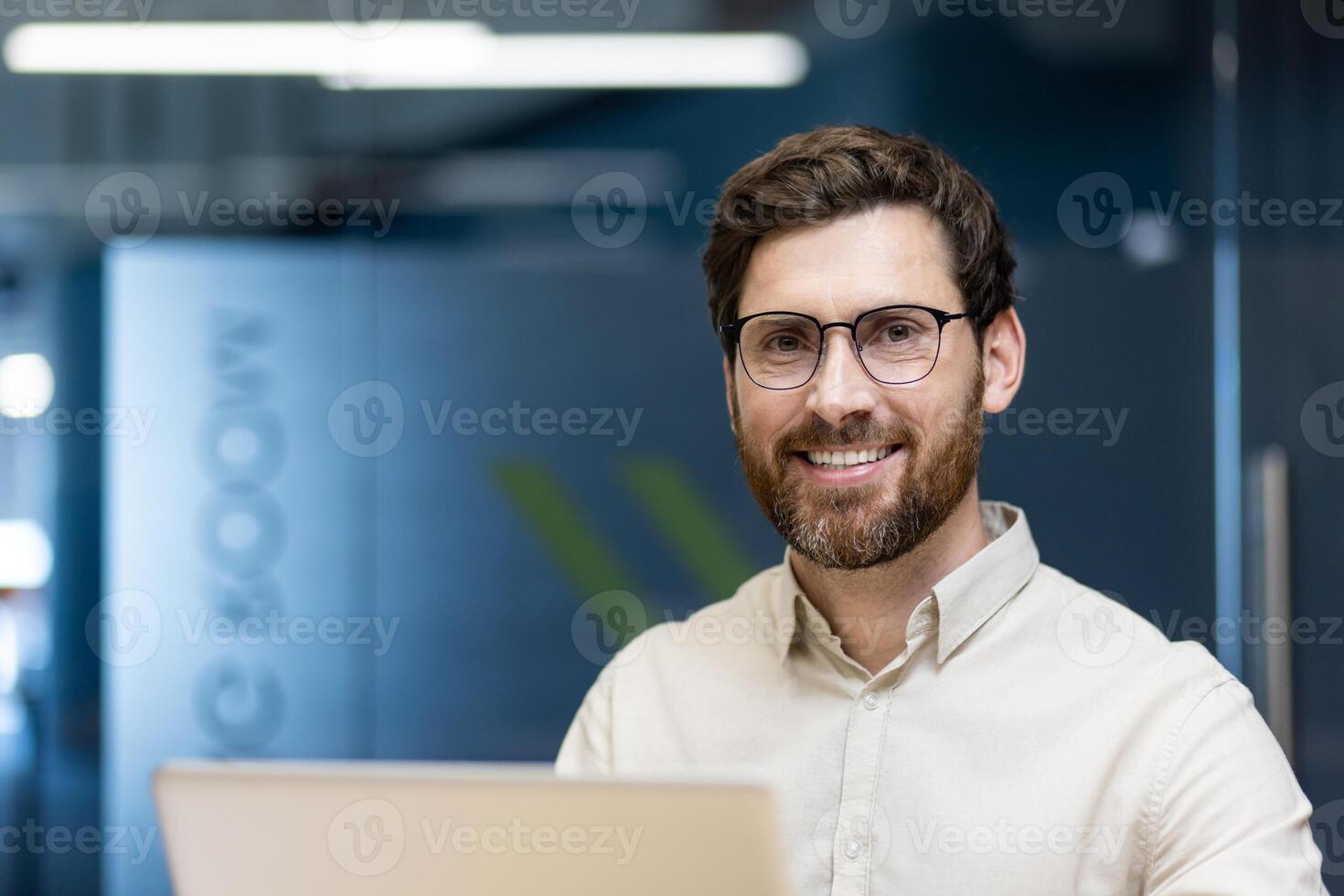 Close-up portrait of a smiling young businessman man in a light shirt and glasses sitting in the office at a laptop and looking at the camera. photo